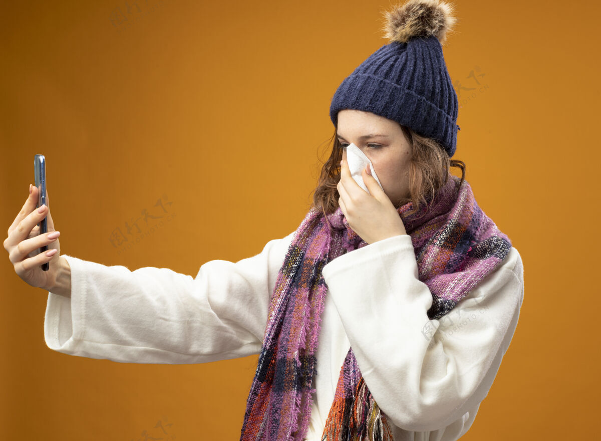 穿穿着白色长袍 戴着带围巾的冬帽 自拍 用隔离在橙色墙上的餐巾擦鼻子的虚弱的生病少女疾病冬天湿巾