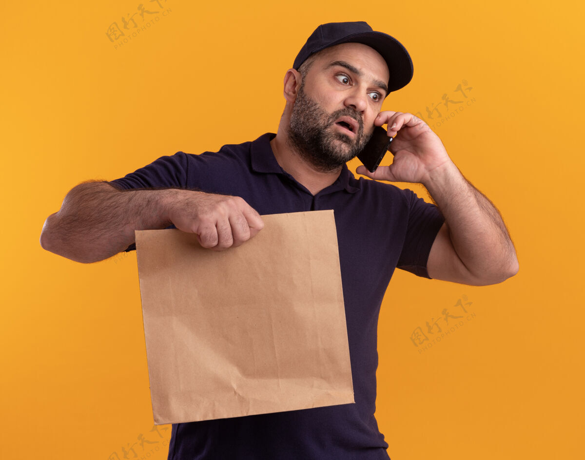 拿着惊恐地看着身边穿着制服 戴着帽子的中年送货员拿着纸食品包讲着电话隔离在黄色的墙上包装男人说话