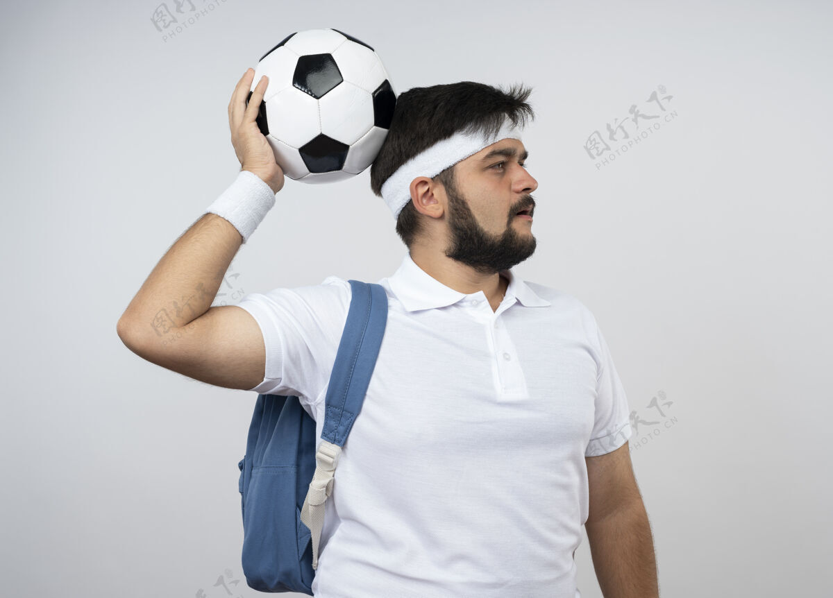 思考一边戴着头带 一边戴着手环 背着背包 一边思考着年轻的运动型男人 一边把球放在隔离在白色墙壁上的头上年轻球背包