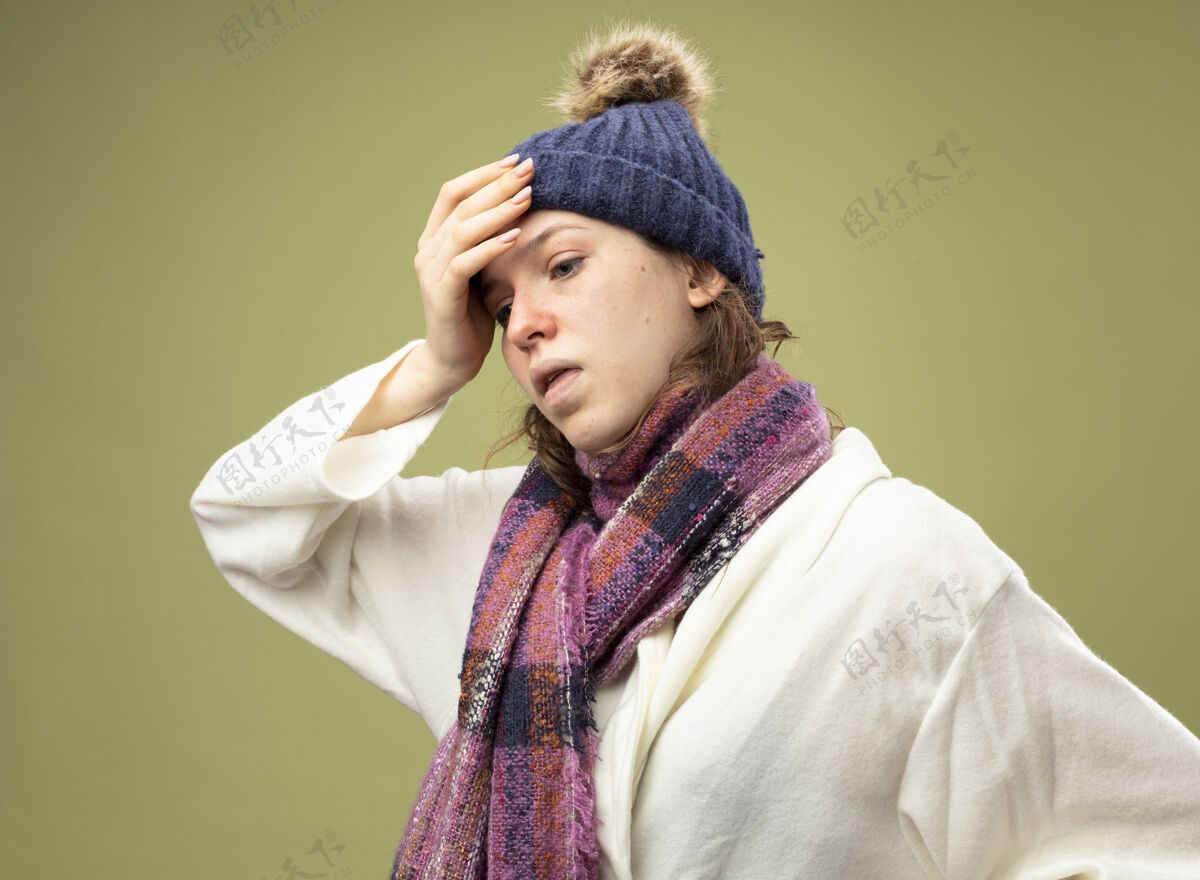 疾病咳嗽的年轻生病的女孩一边看一边穿着白色长袍 戴着冬天的帽子 戴着围巾 手放在额头上 隔离在橄榄绿上额头年轻围巾