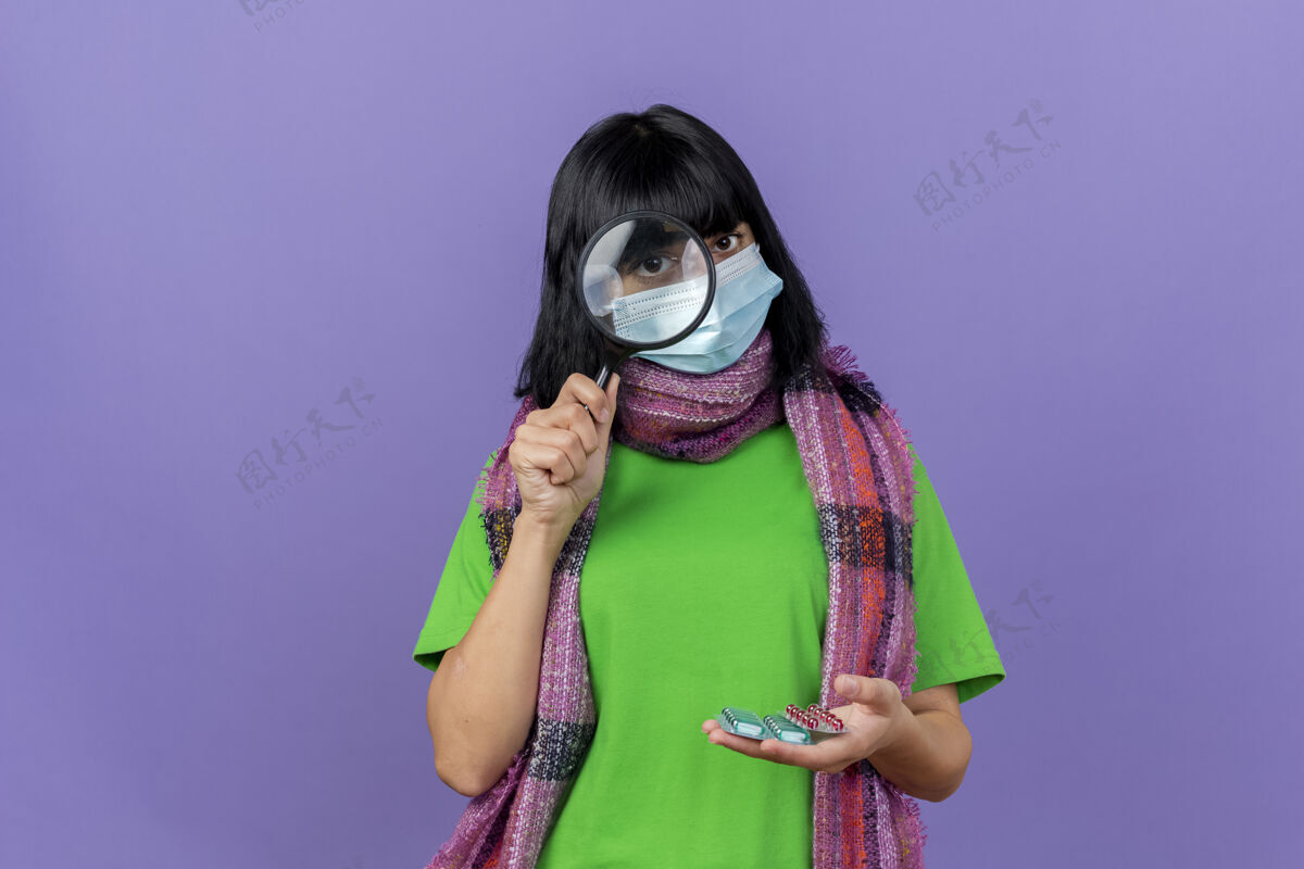抱着戴着口罩和围巾的年轻病妇拿着一包包胶囊透过放大镜看前面 隔离在紫色的墙上 留着复印空间包穿年轻人