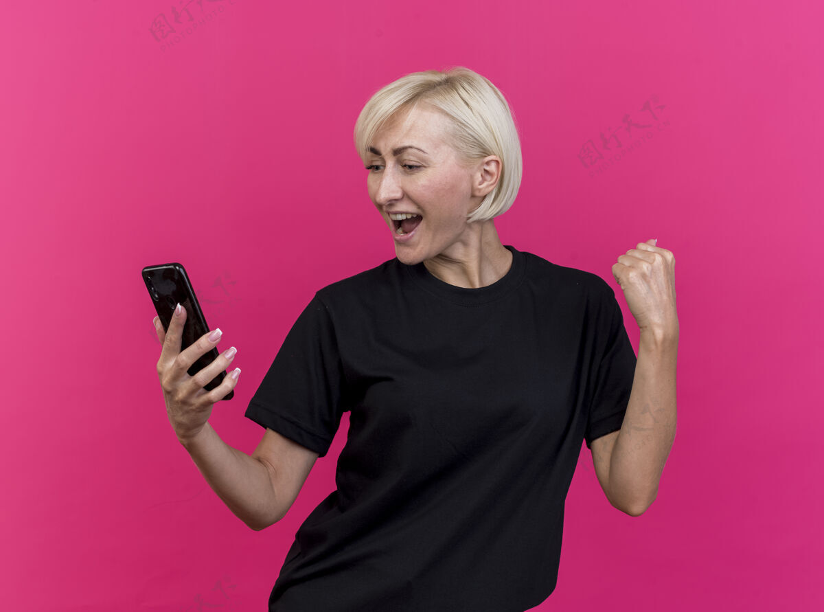 感觉快乐的中年金发斯拉夫女人拿着手机看着做是的手势孤立在粉红色的墙上衣服市民手机