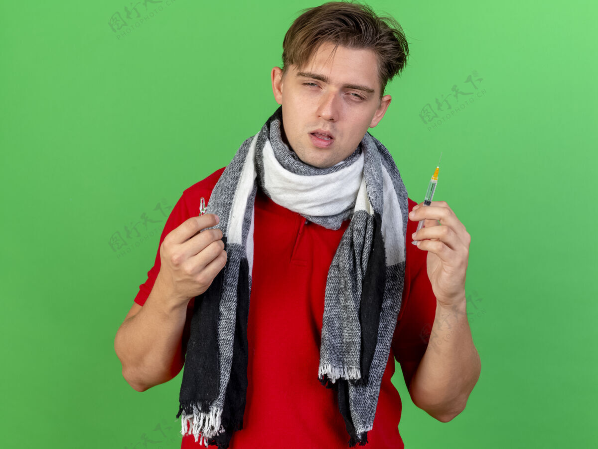 感情虚弱的年轻英俊的金发病男子戴着围巾拿着注射器和医疗安瓿看着隔离在绿色墙上的前面人虚弱金发