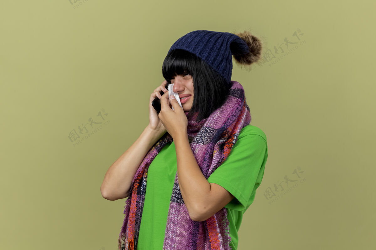 悲伤戴着冬日帽子 戴着围巾的悲伤的年轻病妇站在侧视图里讲着电话看着笔直地擦着眼泪隔离在橄榄绿的墙上留着复印空间直女孩人