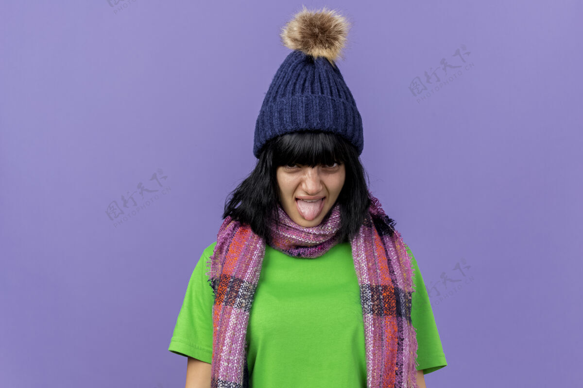 疾病愁眉不展的年轻病女人戴着冬天的帽子和围巾 看着前面显示舌头孤立在紫色的墙壁与复制空间立场姿势围巾