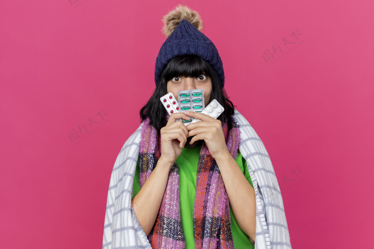 嘴印象深刻的年轻生病的女人戴着冬天的帽子和围巾包裹在格子布手里拿着药片包在嘴前看着前面孤立的粉红色墙壁上的复制空间前面药丸人
