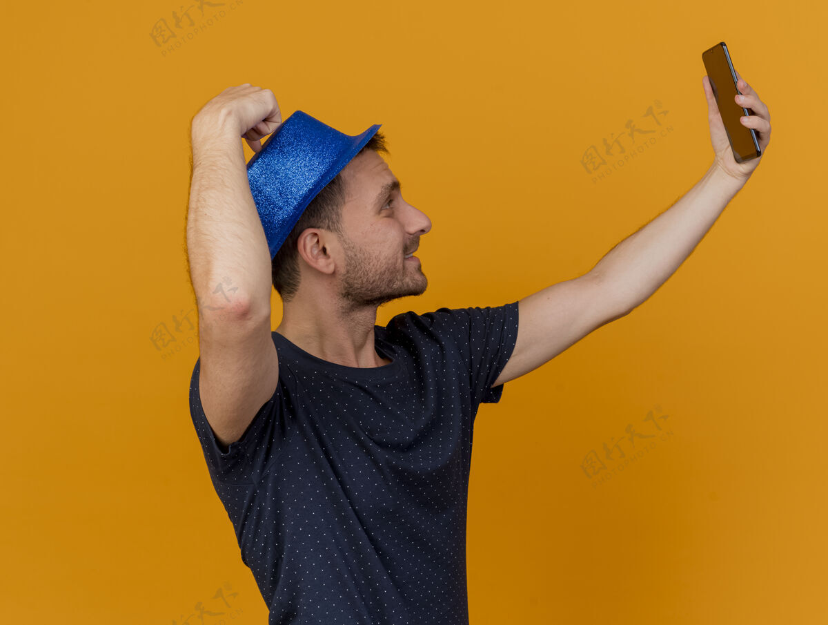 人戴着蓝色派对帽的帅哥高兴地站在一边自拍 看着橘色墙上的手机 还有复制空间站着帽子聚会