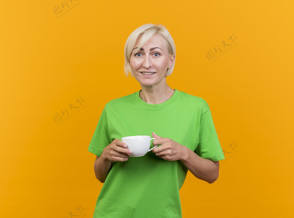 女人微笑的中年金发斯拉夫妇女端着一杯茶 看着前面隔离的黄色墙壁上的复制空间人姿势茶