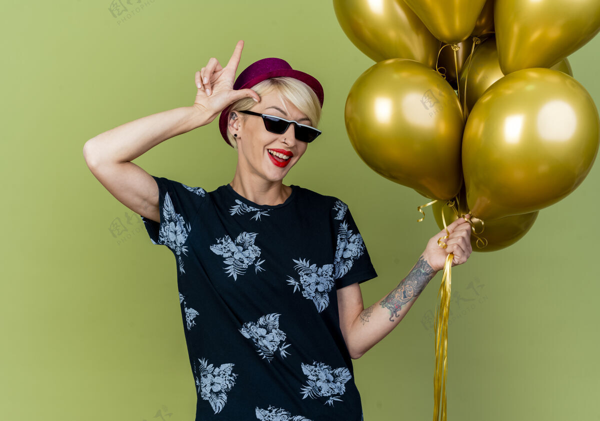 立场快乐的年轻金发派对女郎戴着派对帽和太阳镜举着气球在橄榄绿的墙上做着失败者的手势人人气球