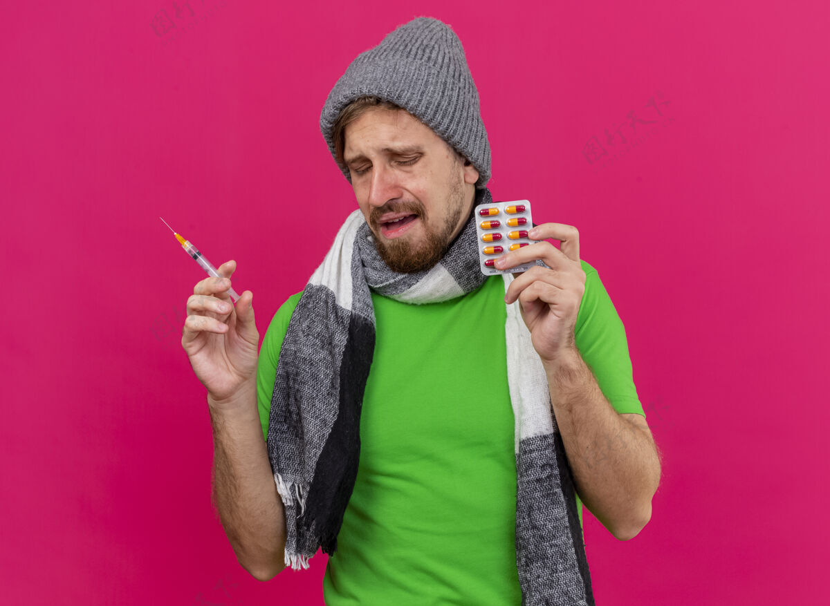 人悲伤的年轻英俊的斯拉夫病夫 戴着冬天的帽子 戴着围巾 手里拿着注射器和一包胶囊 闭着眼睛 隔离在粉红色的墙上 留着复印空间脸疾病帽子