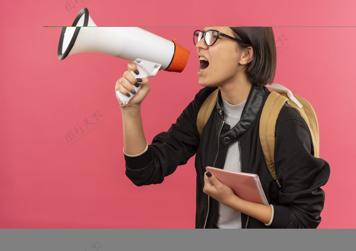 扬声器戴着眼镜和背包的年轻女学生站在侧视图中 拿着便笺簿 由隔离在粉色背景上的演讲者讲话站抱包