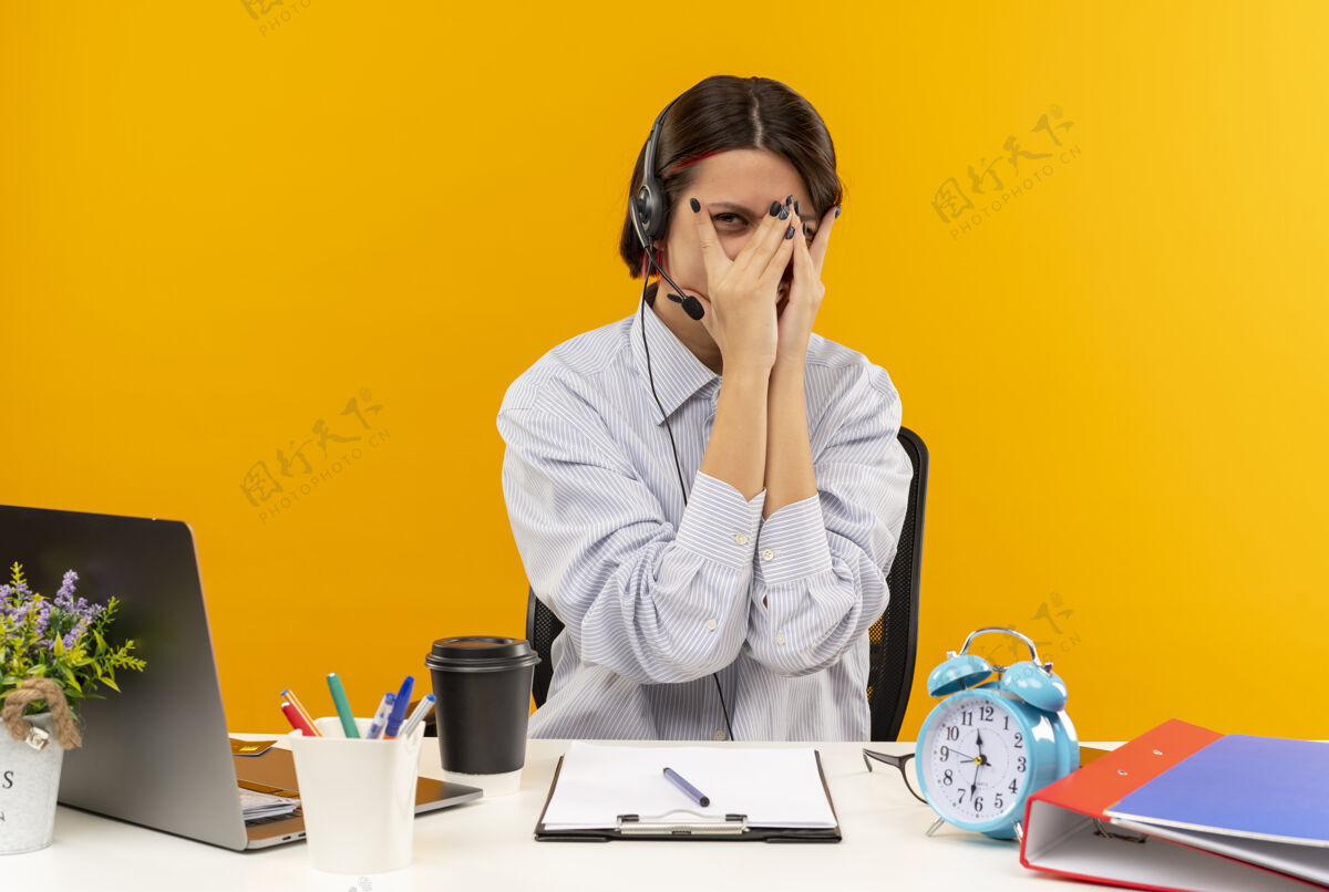 相机年轻的呼叫中心女孩戴着耳机坐在办公桌旁 用工作工具捂住脸 用手指隔着橙色背景看着摄像机手盖工作