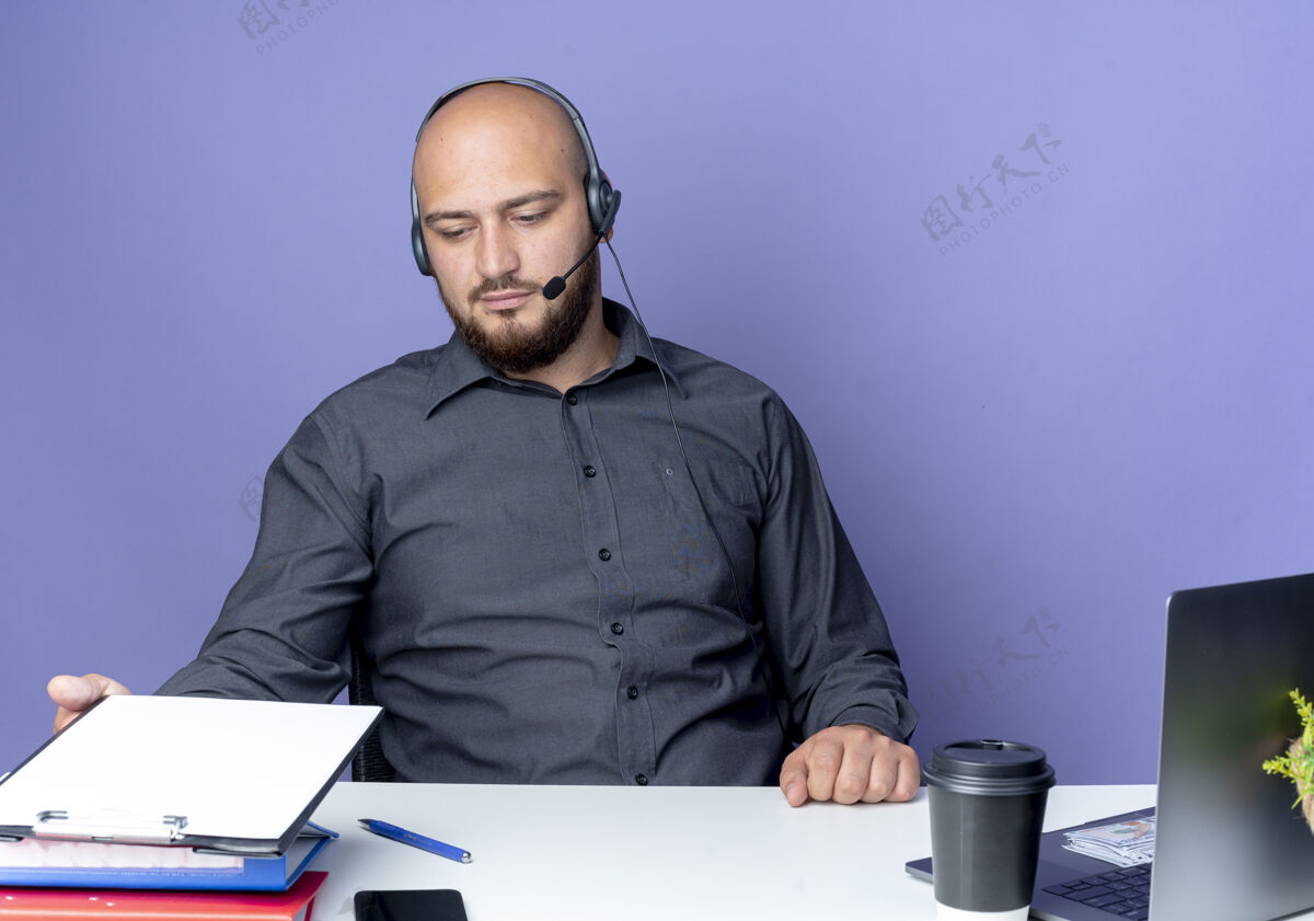 秃头年轻秃顶的呼叫中心男子戴着耳机坐在办公桌旁 拿着工作工具抚摸着 看着紫色背景上孤立的剪贴板工作年轻中心
