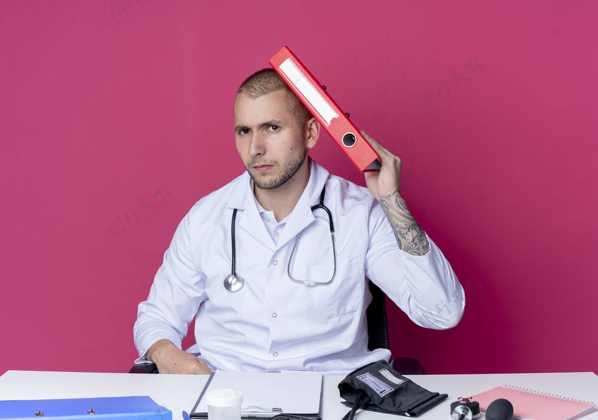 穿着穿着医用长袍和听诊器的年轻男性医生坐在办公桌旁 工作工具接触着头部 文件夹隔离在粉色背景上坐着年轻头