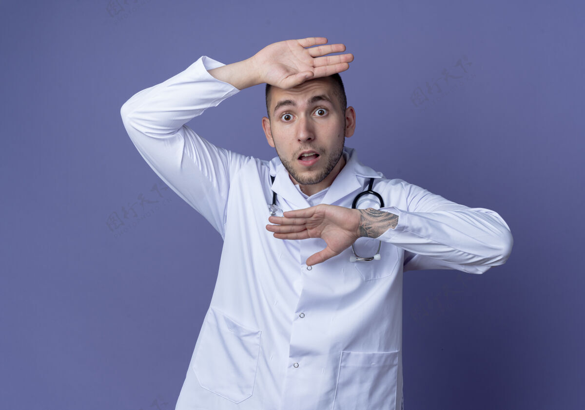 长袍印象深刻的年轻男医生穿着医用长袍 脖子上戴着听诊器 手放在头上 头下隔离在紫色上医疗紫色听诊器