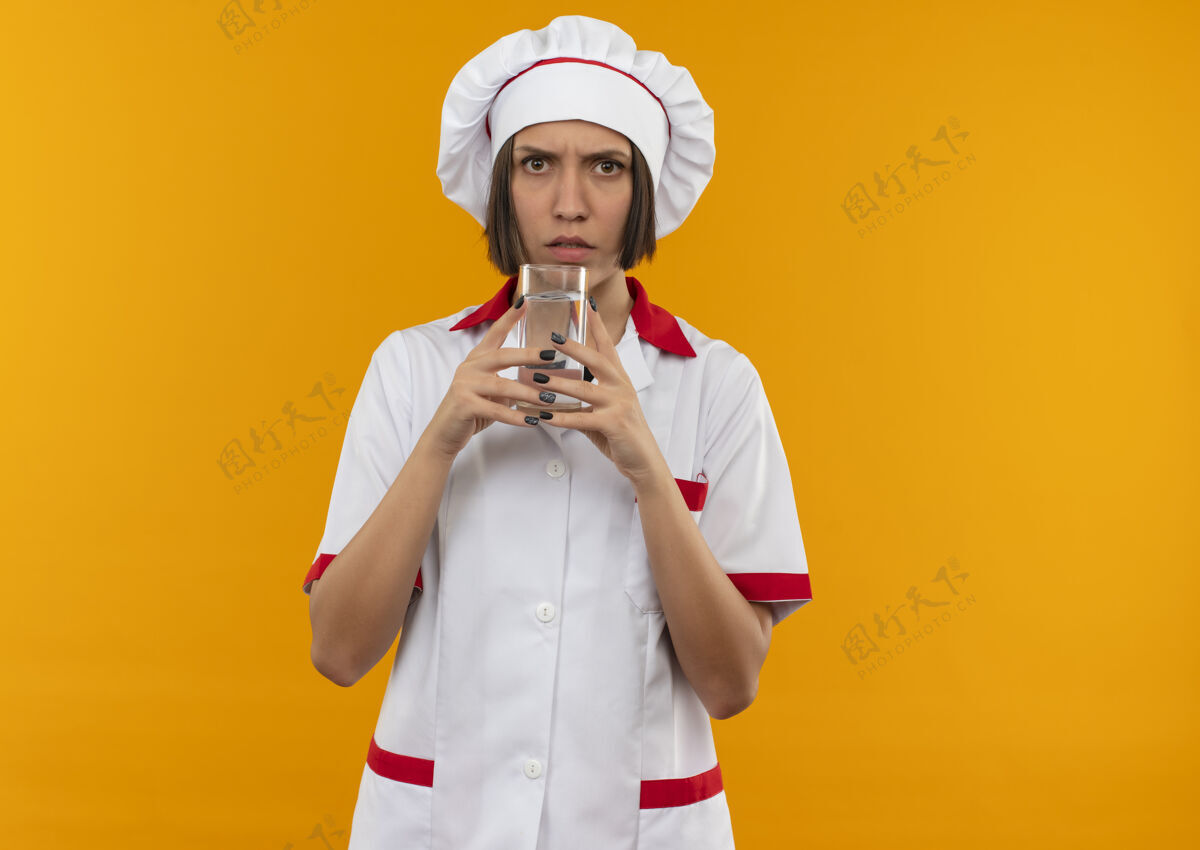 年轻穿着厨师制服的年轻女厨师拿着一杯水 看着隔离在橙色背景上的相机 还有复印空间烹饪女拿着