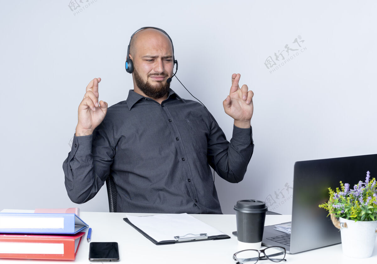耳机戴着耳机的秃头呼叫中心年轻人坐在办公桌旁 手里拿着工作工具 交叉着手指看着笔记本电脑 背景是白色的工作工具交叉