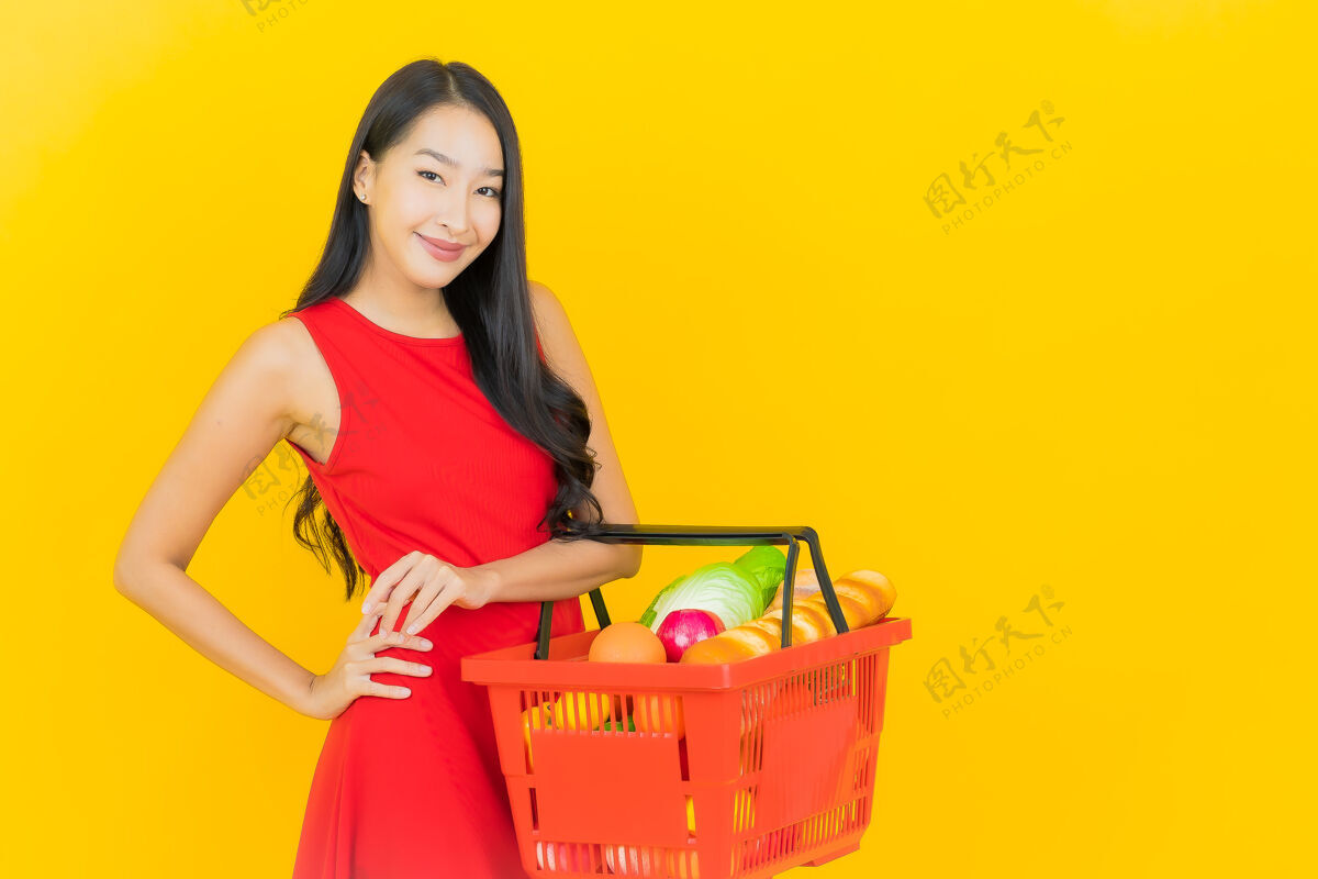 持有一幅美丽的亚洲年轻女子微笑的画像 黄色的墙上挂着超市的购物篮购买微笑商场