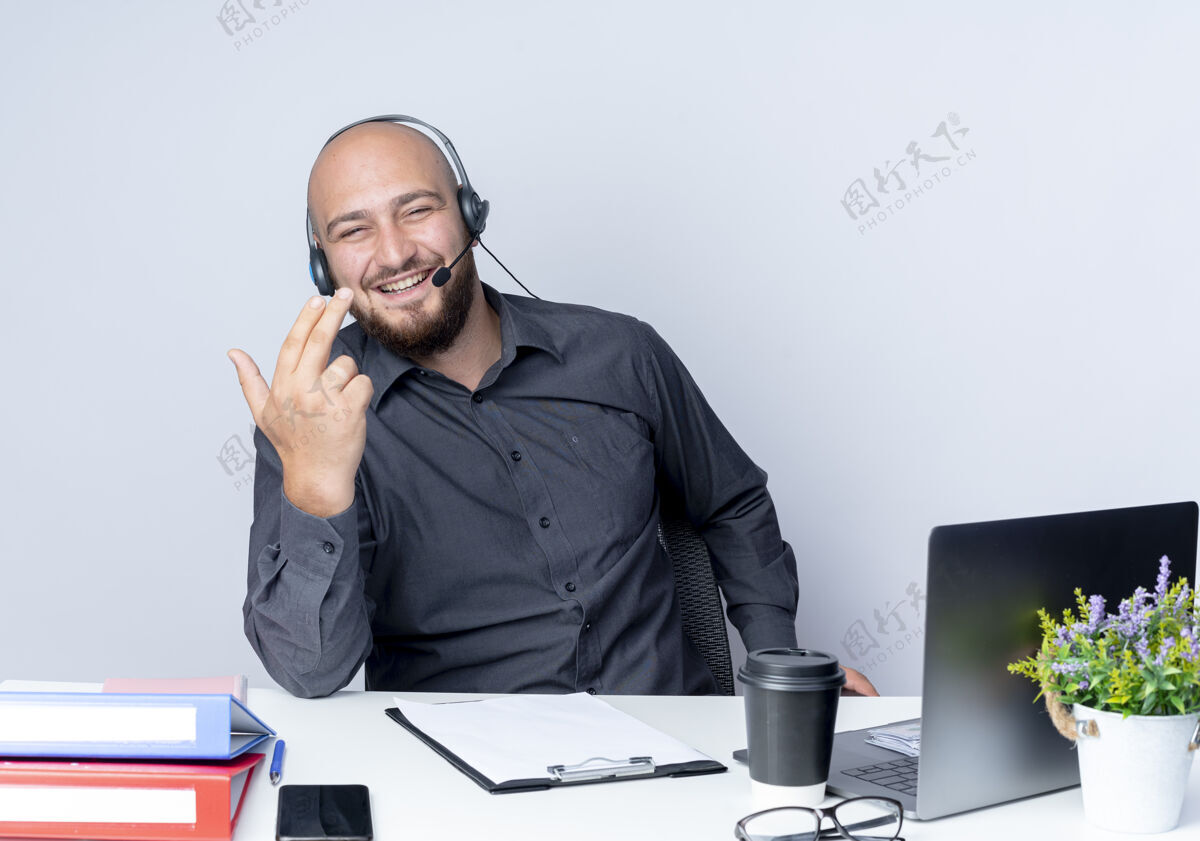 坐着快乐的年轻秃头呼叫中心男子戴着耳机坐在办公桌旁 手上拿着工作工具做手枪手势 手上隔离着白色耳机工作工具