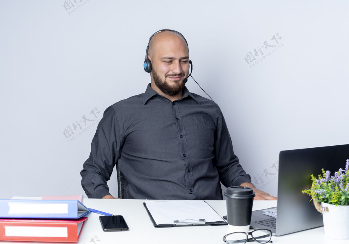 工具高兴的年轻秃头呼叫中心男子戴着耳机坐在办公桌旁 用工作工具看着隔离在白色背景上的笔记本电脑耳机坐着笔记本电脑