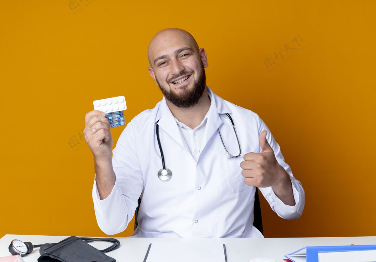 抱着面带微笑的年轻秃顶男医生穿着医用长袍 手持听诊器坐在办公桌旁 手里拿着药片 他的拇指孤立地竖立在橙色背景上年轻人工具穿