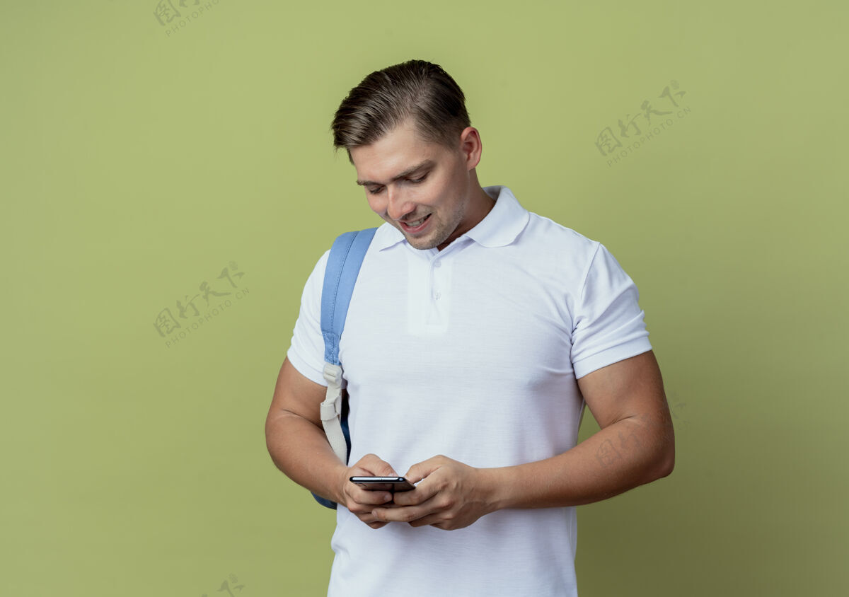 微笑面带微笑的年轻帅气男学生背着书包拿着手机看着橄榄绿背景上的孤立帅气抱着橄榄