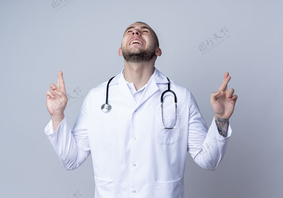 长袍快乐的年轻男医生 穿着医用长袍 脖子上戴着听诊器 闭着眼睛 做着双手交叉的手势周围听诊器闭合