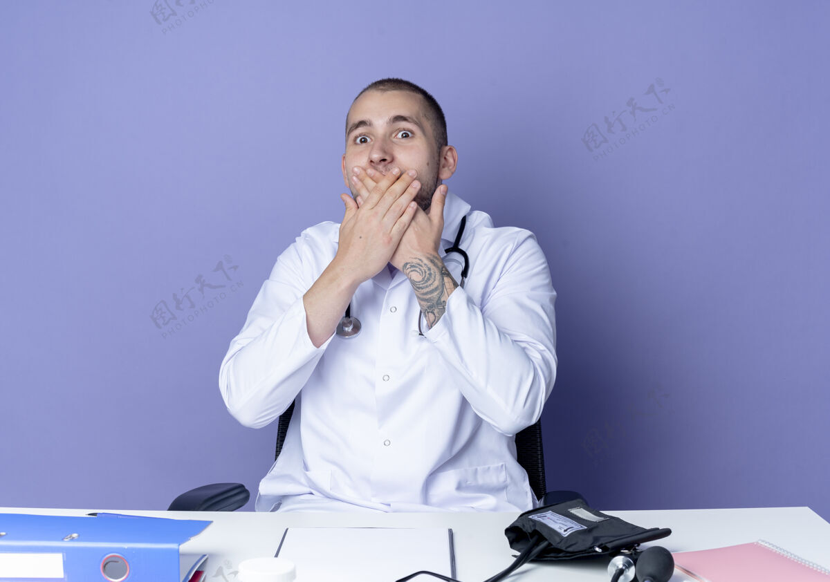 年轻令人印象深刻的年轻男医生穿着医用长袍和听诊器坐在办公桌旁 手放在嘴上 紫色隔离工具穿着医疗