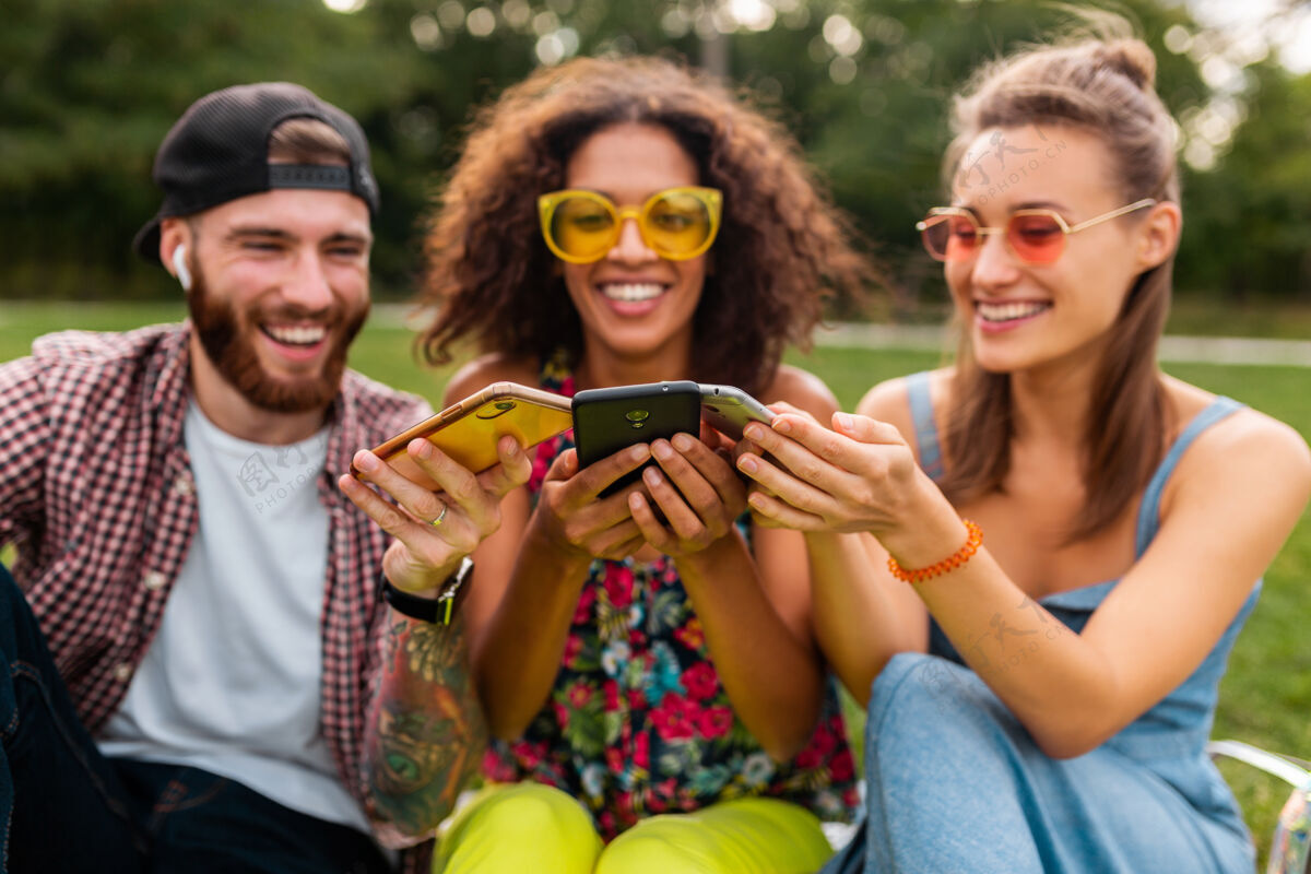 多民族快乐的年轻朋友们坐在公园里用智能手机微笑 男人和女人一起玩积极黑色享受