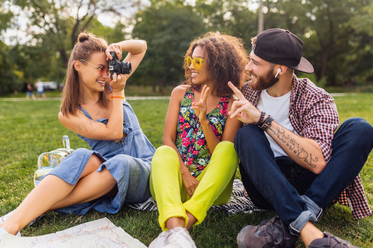多种族快乐的年轻朋友们一起坐在公园里 男女一起玩 带着相机旅行 拍照兴奋人时尚