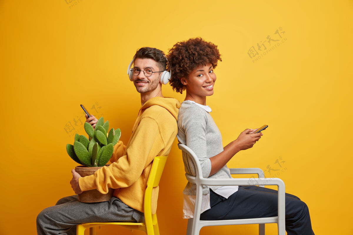 女性不同种族的朋友在椅子上对着鲜艳的黄色墙壁摆姿势 拿着手机 表情愉快地看着手机关系帅哥