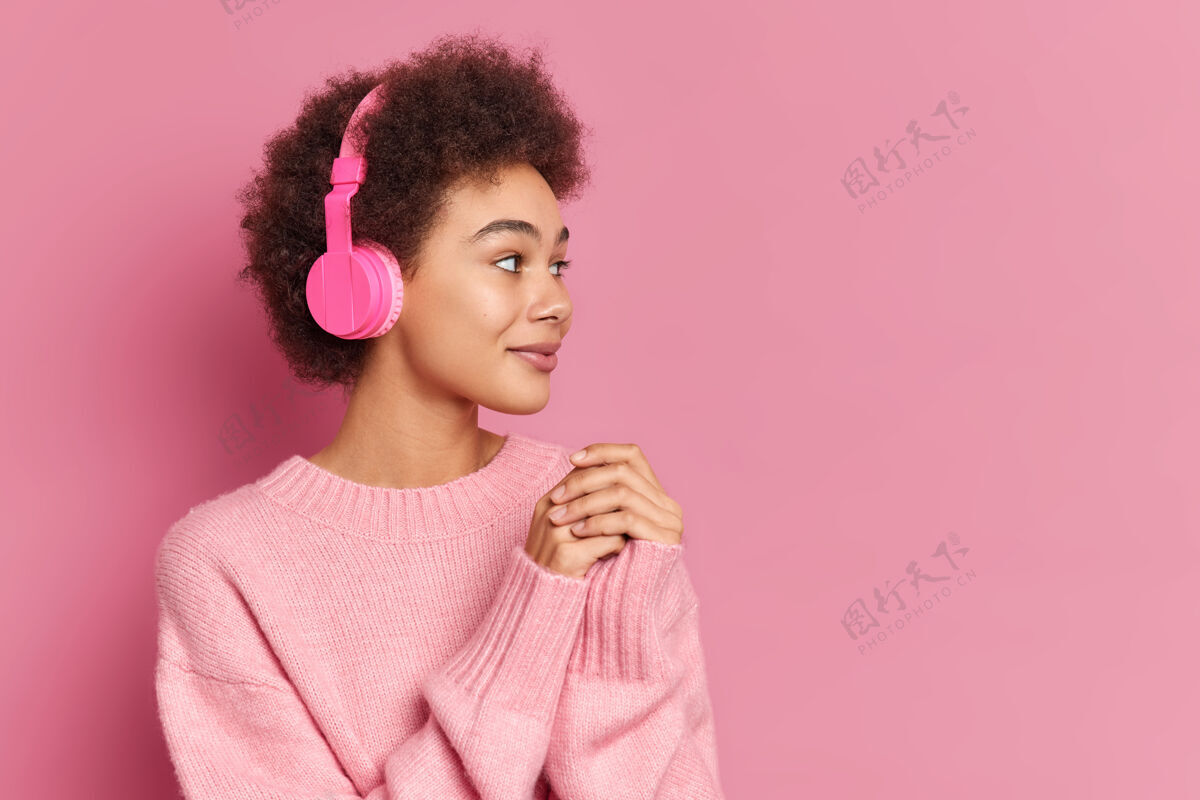 集中漂亮的卷发民族女子戴着立体声耳机 专注在一边听音乐休闲套头衫姿势粉色模型