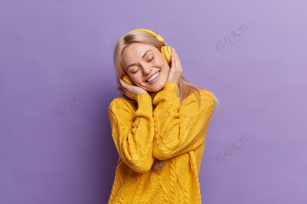 活跃积极的金发欧洲女人歪着头微笑睁大眼睛享受每一点音乐戴着无线耳机穿着黄色毛衣乐观紫色漂亮