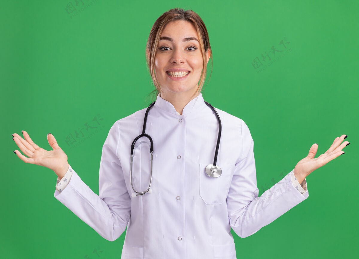 手面带微笑的年轻女医生穿着医用长袍 听诊器摊开双手隔离在绿色的墙上传播听诊器医疗