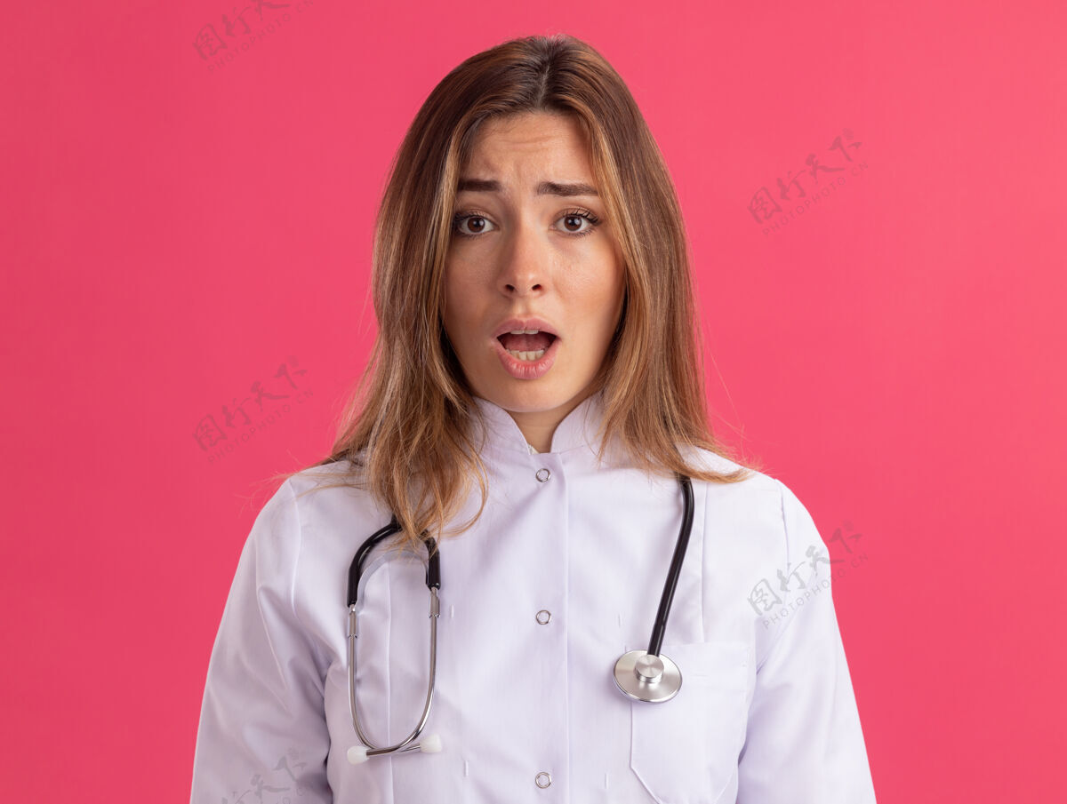 医疗困惑地看着镜头年轻的女医生穿着医用长袍听诊器隔离在粉红色的墙上年轻女性听诊器