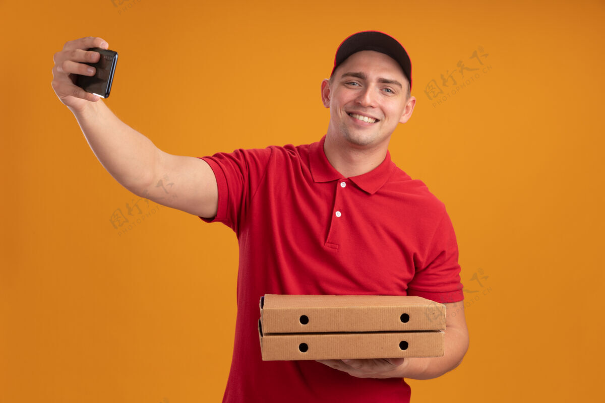 拿着微笑的年轻送货员穿着制服 戴着帽子 拿着披萨盒 在橙色的墙上自拍制服盒子帽子