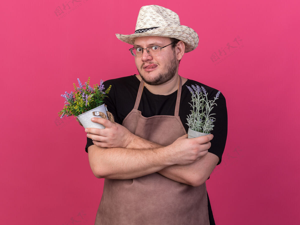 穿可疑的年轻男园丁戴着园艺帽 拿着和粉红色墙上隔离的花盆里的花交叉交叉帽子可疑