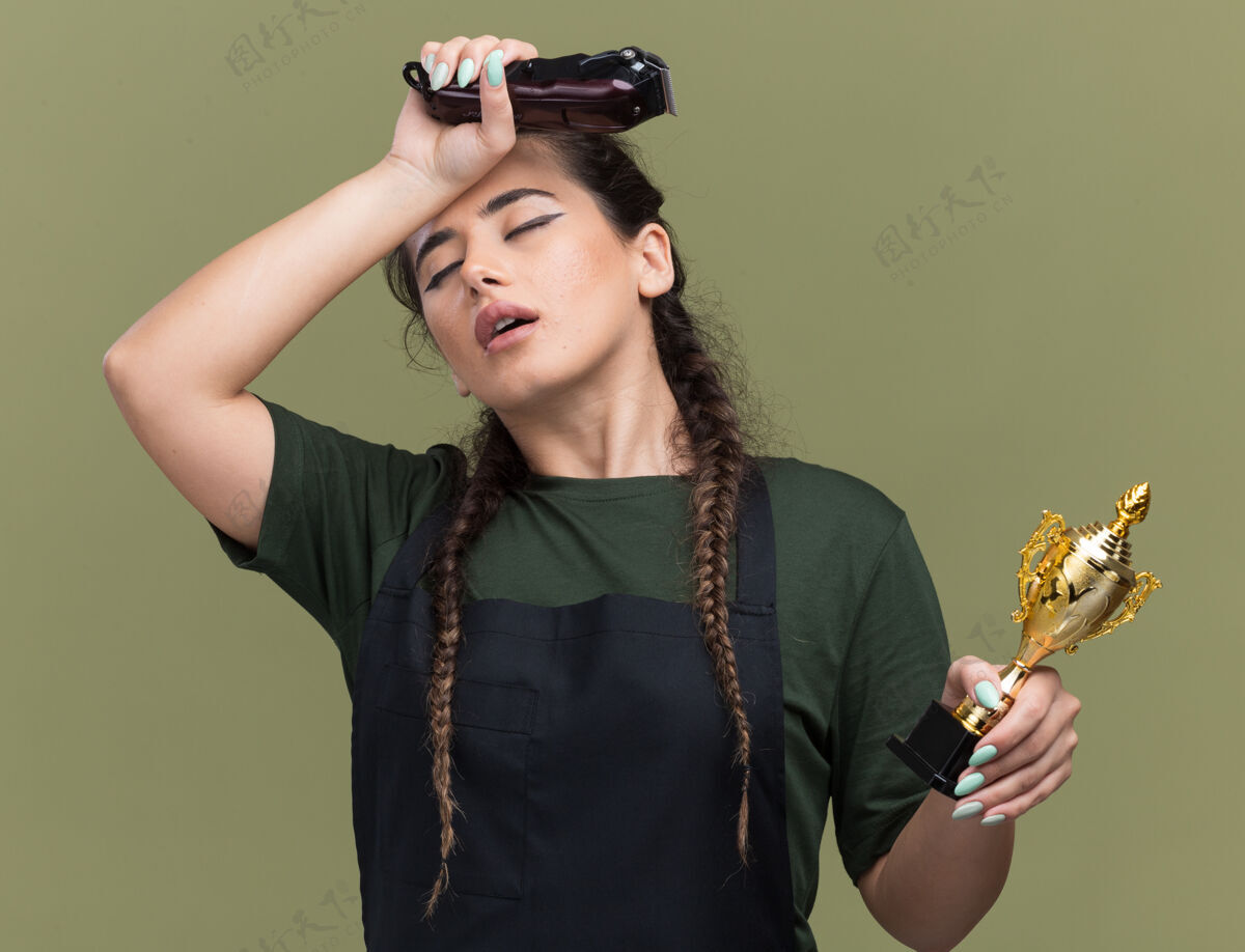 杯子厌倦了闭着眼睛年轻的女理发师穿着制服拿着理发器拿着优胜者杯把手放在额头上隔离在橄榄绿的墙上制服持有女