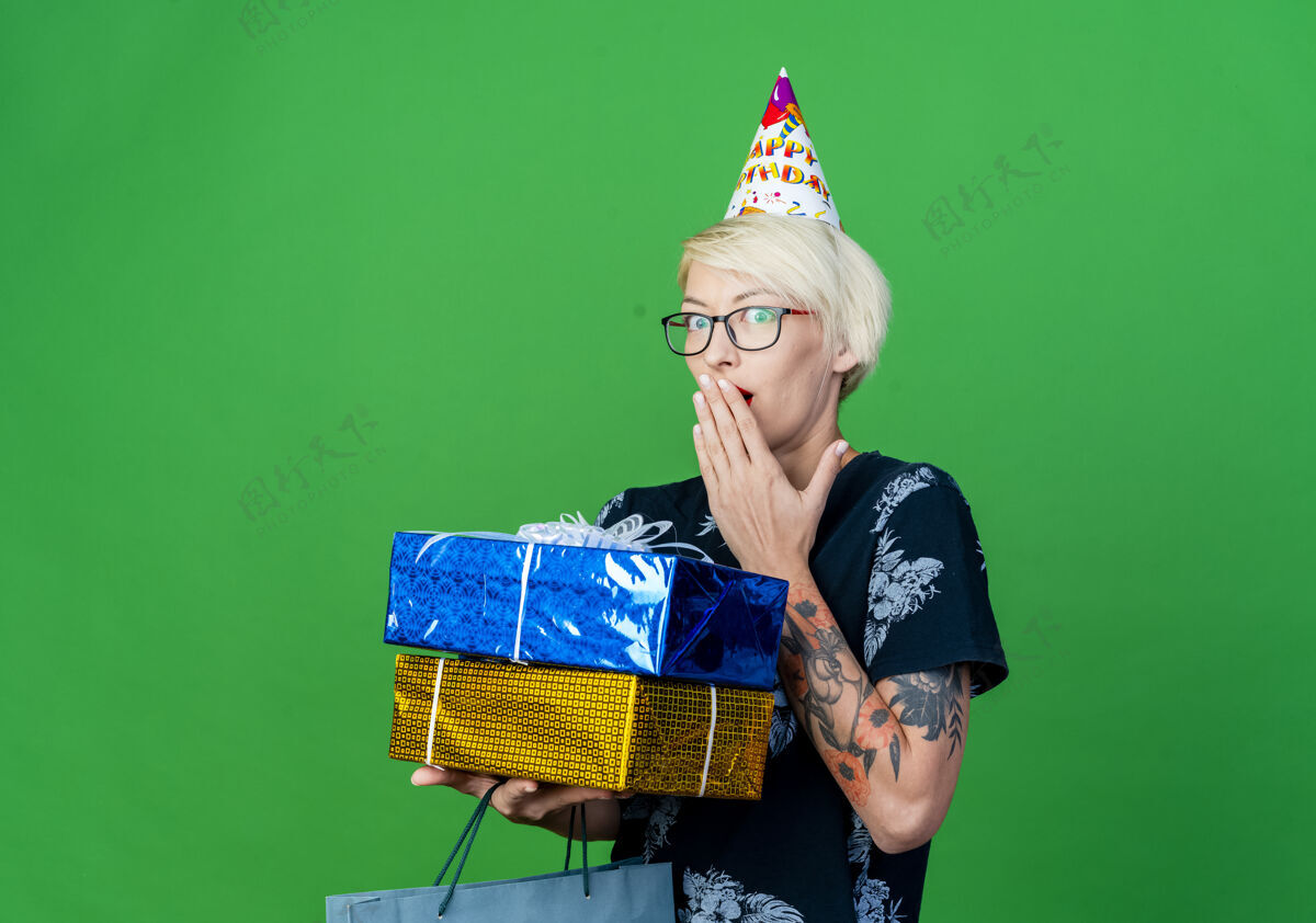 嘴惊讶的年轻金发派对女孩戴着眼镜 戴着生日帽 拿着纸袋和礼品盒 看着相机 手放在嘴上 在绿色的背景上与复印空间隔离年轻绿色生日