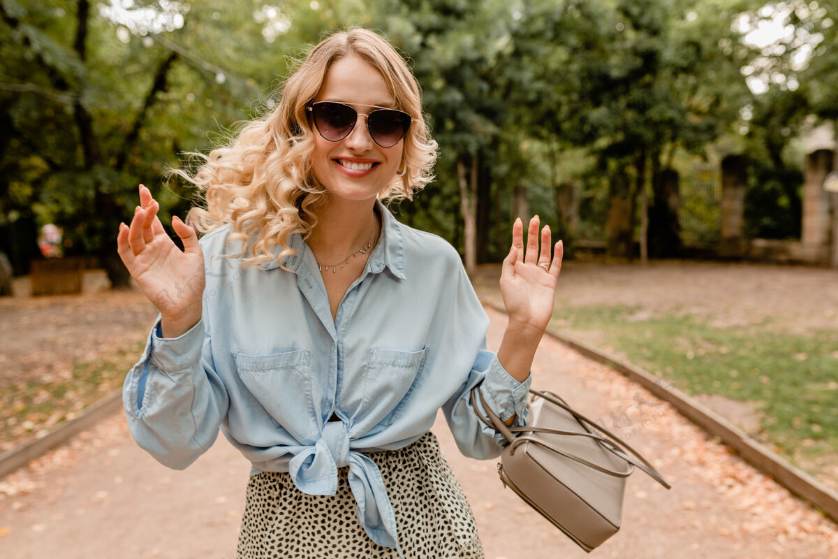 手提包迷人的金发美女穿着时髦的服装 戴着优雅的太阳镜和钱包在公园散步时尚时尚优雅