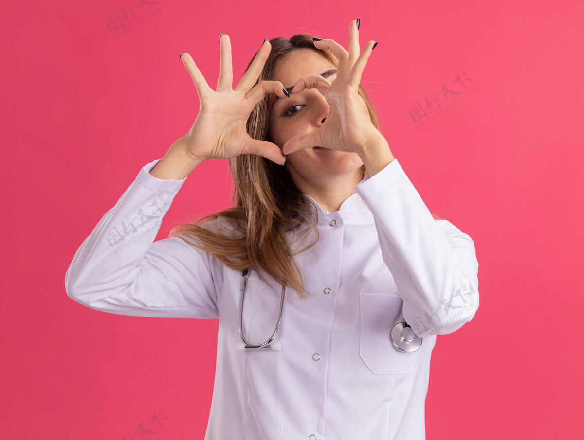 女性年轻的女医生穿着医用长袍 听诊器显示出孤立在粉红色墙上的心脏姿势长袍表演手势