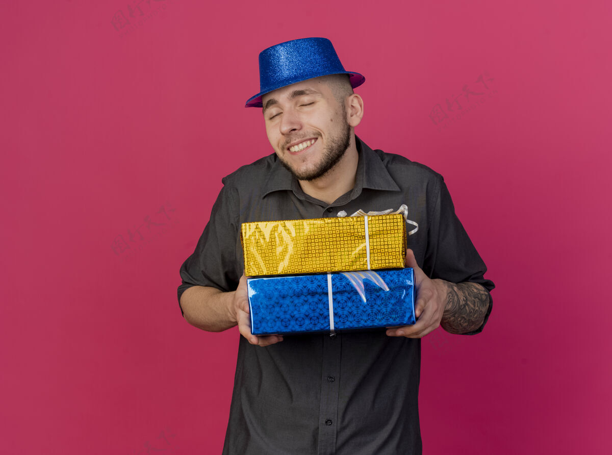 帽子面带微笑的年轻帅哥斯拉夫党人戴着党帽拿着礼品包闭上眼睛孤立在深红色背景与复制空间男人礼物眼睛