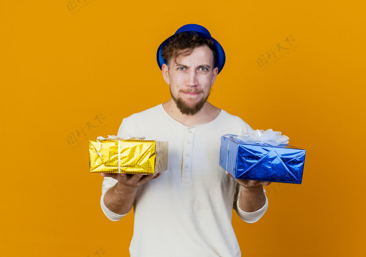 聚会困惑的年轻英俊的斯拉夫党人戴着党的帽子拿着礼品盒看着相机孤立的橙色背景与复制空间橙色帽子年轻