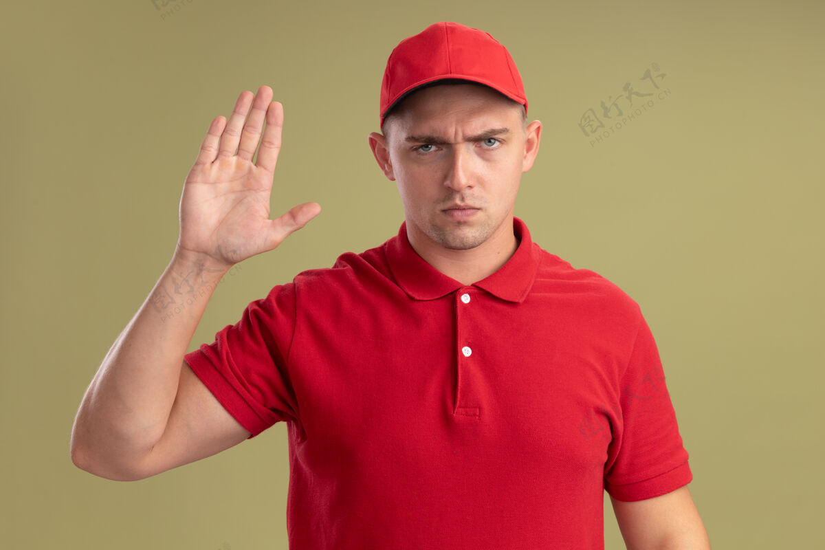 年轻严格的年轻送货员穿着制服 戴着帽子 在橄榄绿的墙上显示停止手势橄榄交货帽子