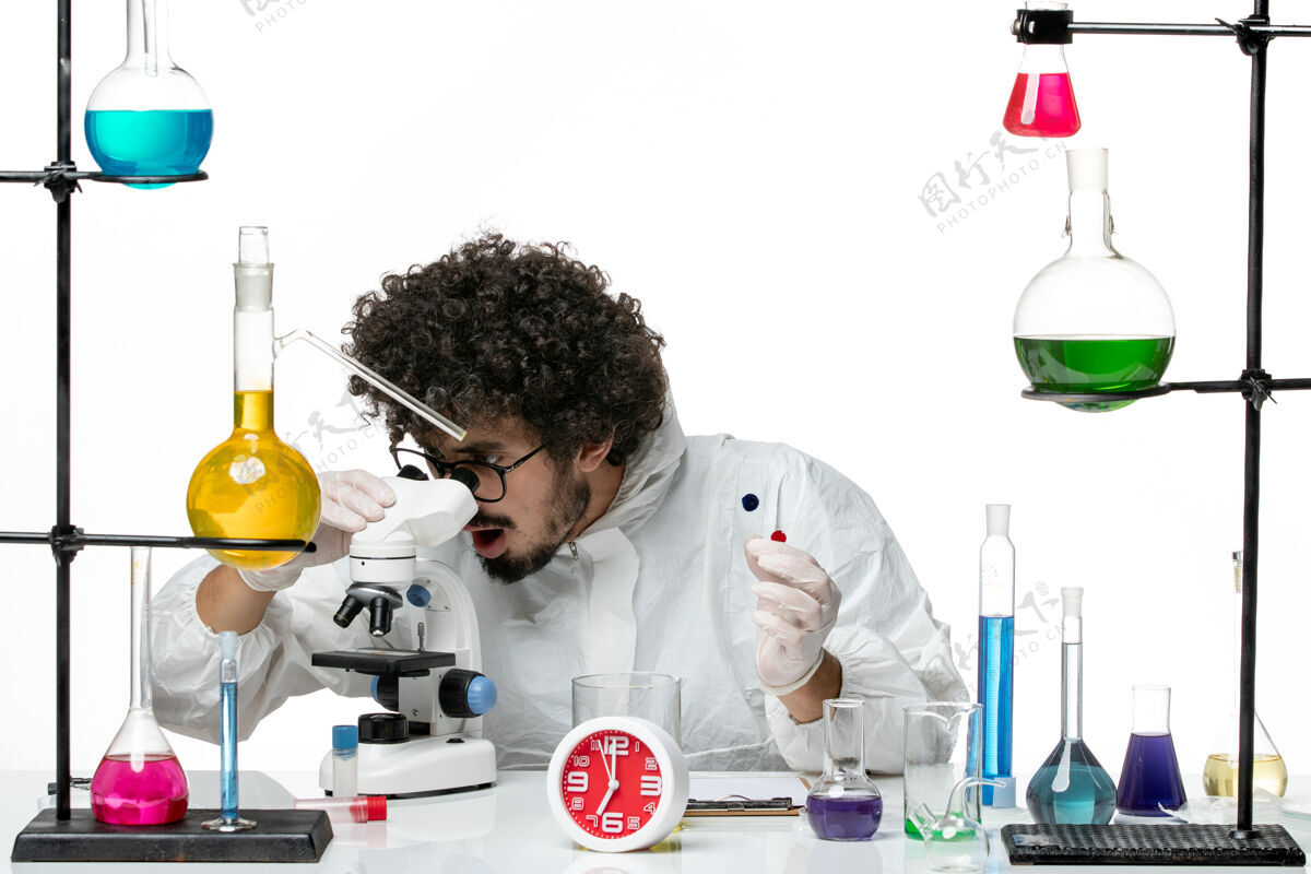 化学正面图身着特殊套装的年轻男科学家拿着样品 在白墙上使用显微镜医学实验室实验室外套
