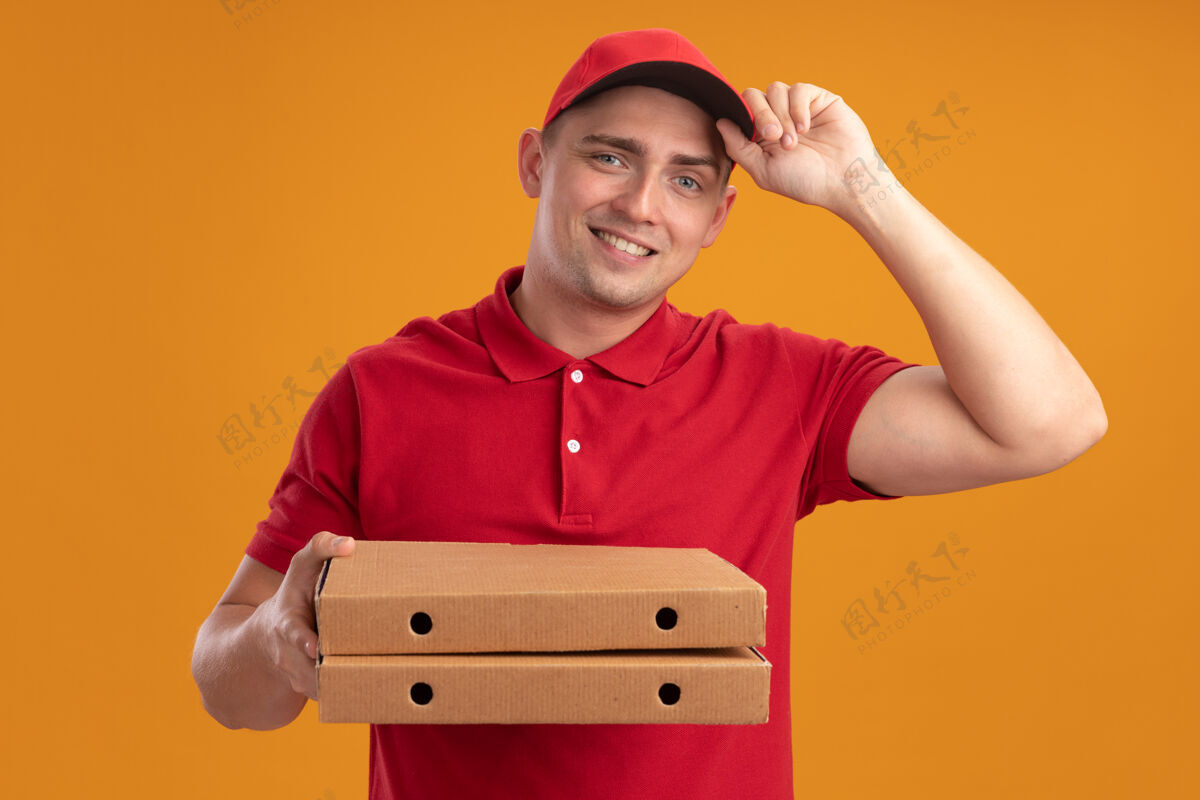 男人微笑着的年轻送货员穿着制服 戴着帽子 拿着隔离在橙色墙上的比萨饼盒送货微笑年轻人