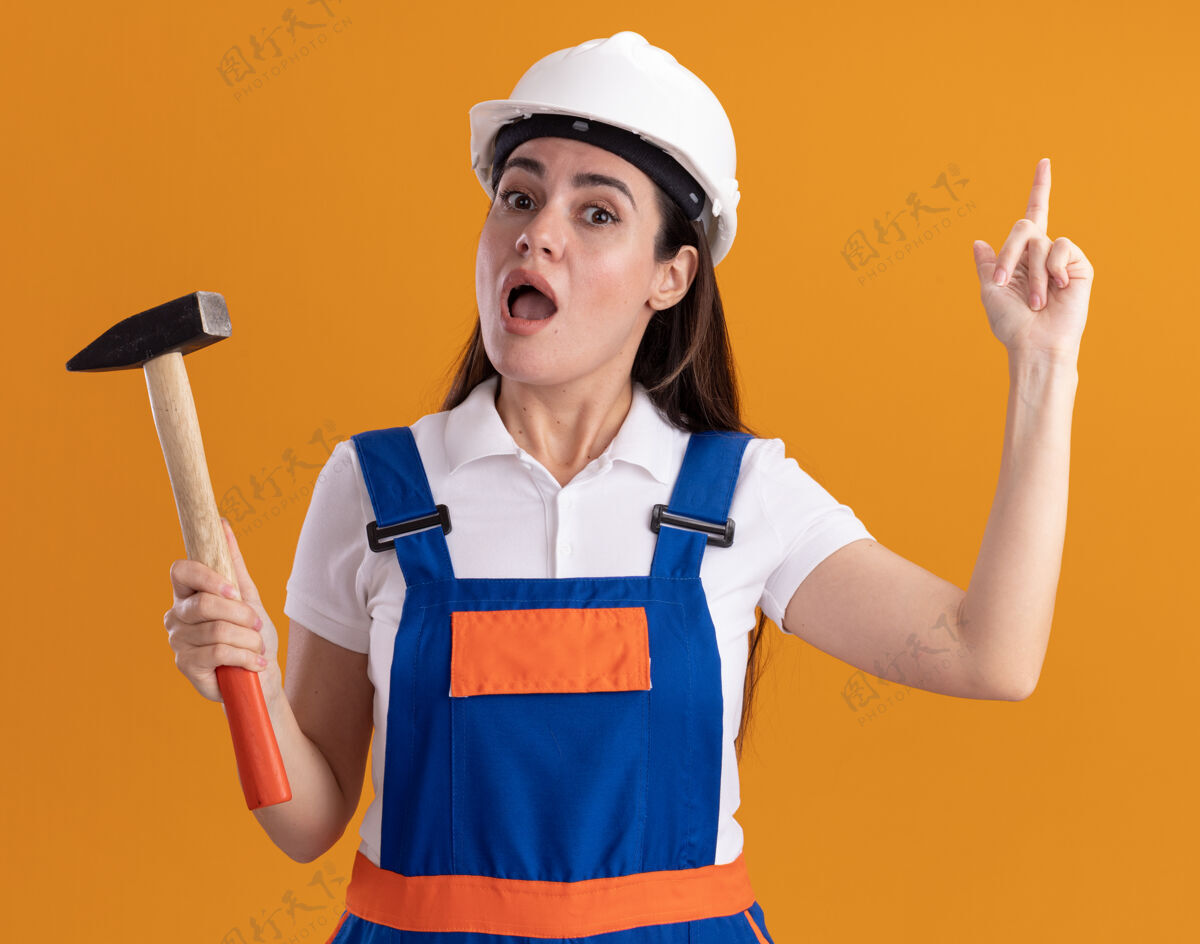 年轻一个穿着制服的年轻建筑女拿着锤子指着隔离在橙色墙上的墙制服持有锤子
