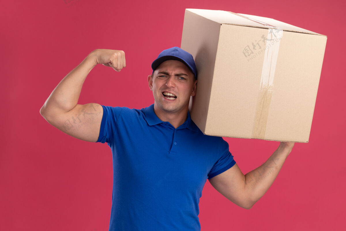 自信自信的年轻送货员穿着制服 戴着帽子 拿着一个大盒子 在粉红色的墙上显示出强烈的姿态穿握着年轻