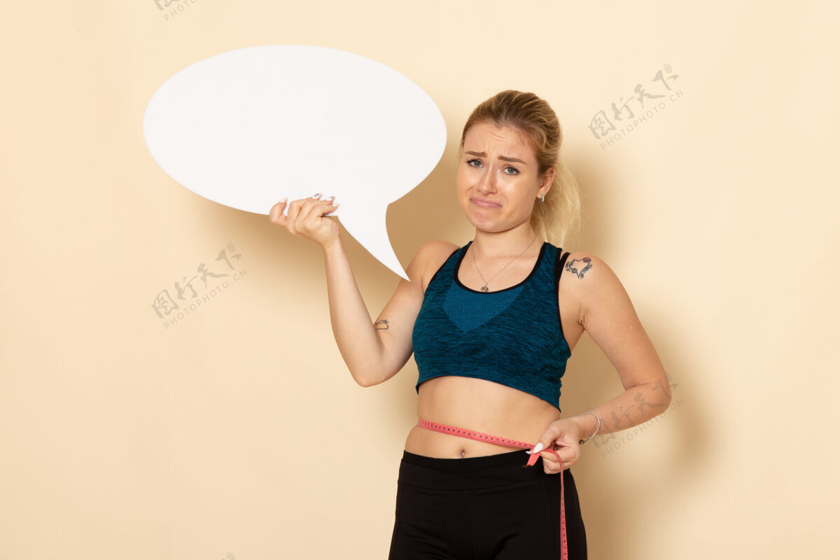 量正面图身穿运动服的年轻女性手持白色标志 在白色墙上测量身体健康运动美容运动适合健身身体腹
