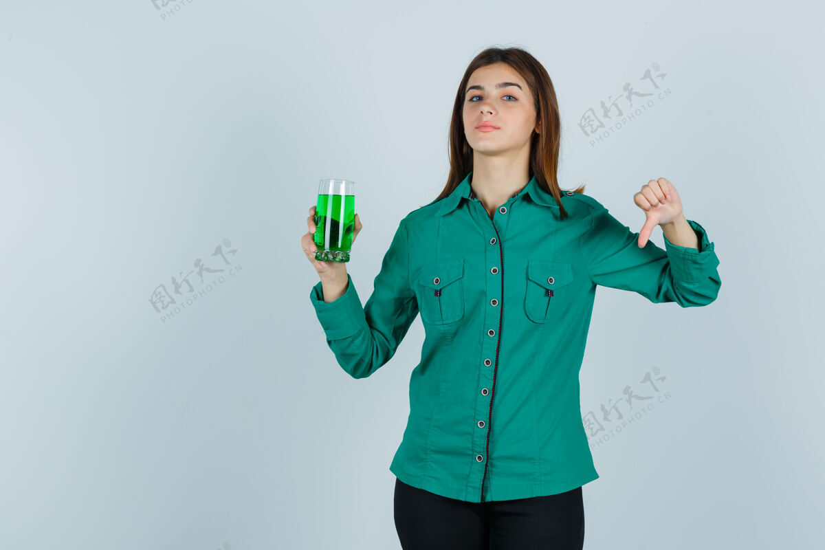 问题年轻女孩拿着一杯绿色液体 穿着绿色上衣 黑色裤子 拇指朝下 看起来很不高兴前视图显示向下前面
